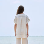 A woman stands with her back to the camera wearing a matching off-white linen two-piece set consisting of a short-sleeve tunic and wide-leg trousers, with thin dark contrast stitching, set against a blurred background of the sea and sky.