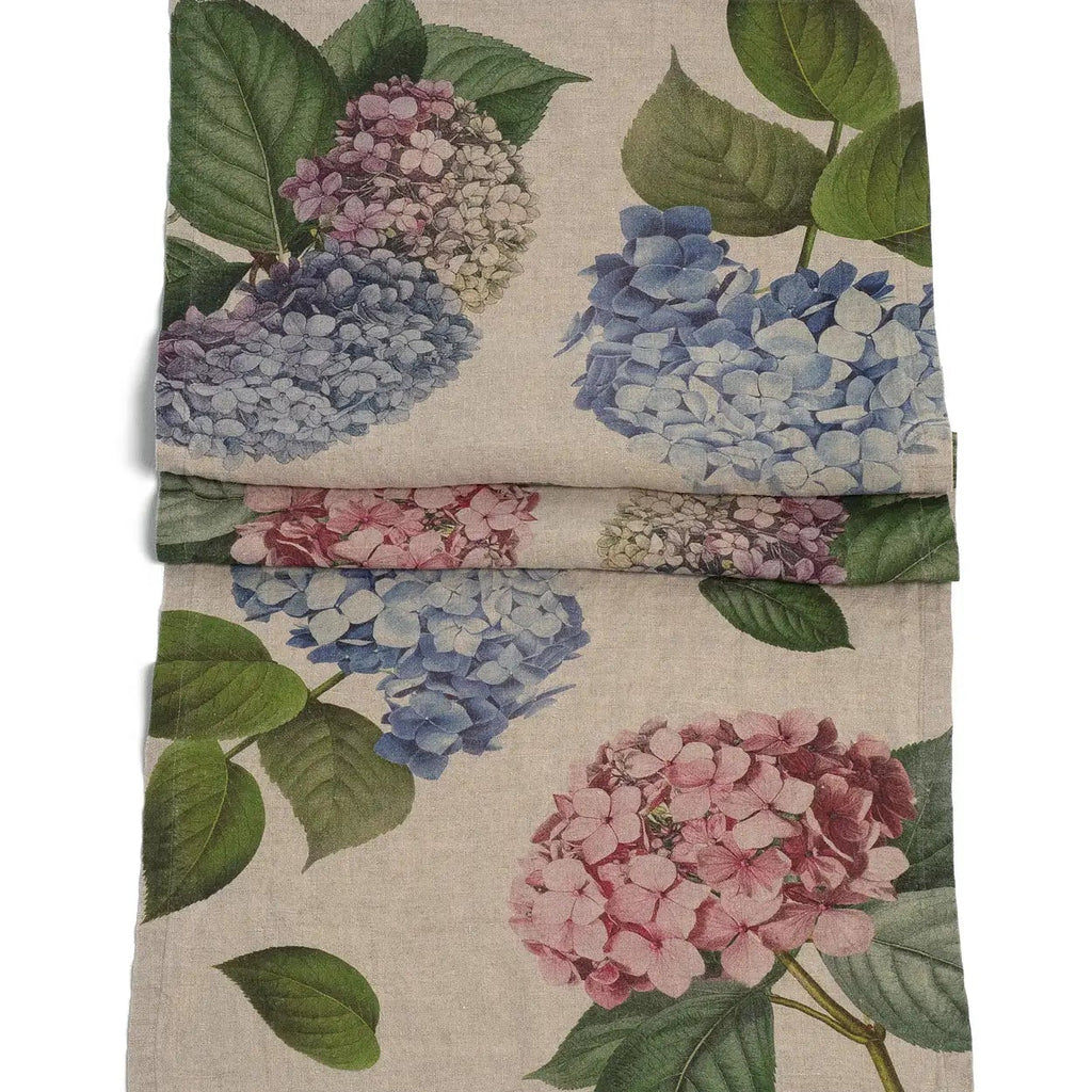 A folded linen napkin featuring a vintage-style botanical print of blue, pink, and lavender hydrangeas with green leaves on a natural beige linen background.
