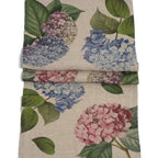 A folded linen napkin featuring a vintage-style botanical print of blue, pink, and lavender hydrangeas with green leaves on a natural beige linen background.