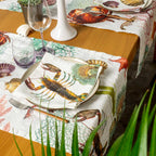 A styled dining table featuring a white table runner and matching place settings with a colorful lobster and seashell print, set on a wooden table with glassware and a white candle holder.