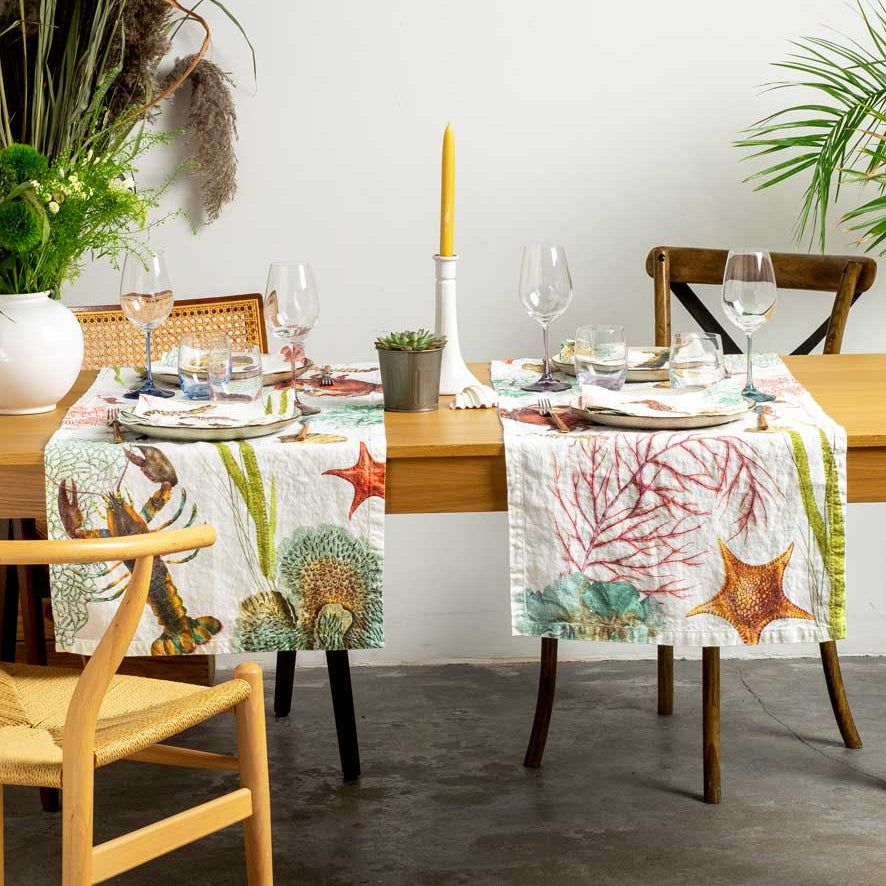 A coastal-themed table runner set on a wooden dining table, featuring a main runner and matching placemats with starfish, sea urchins, and coral patterns, styled in a bright dining room.