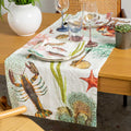 A table runner with a colorful ocean-themed print featuring lobsters, starfish, and sea plants, styled on a wooden dining table with plates, wine glasses, and chairs.