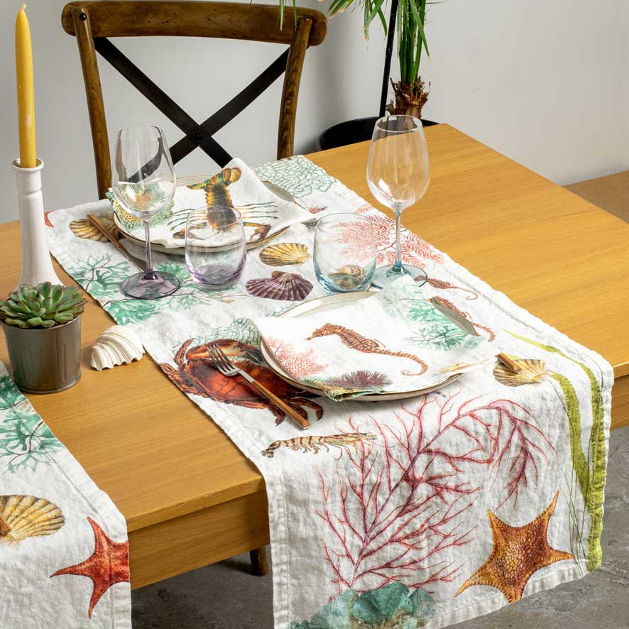 A white table runner with a colorful seashell and starfish print, styled on a wooden dining table with plates, wine glasses, and a candle.