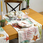 A white table runner with a colorful seashell and starfish print, styled on a wooden dining table with plates, wine glasses, and a candle.