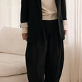 Woman wearing a black coat, beige sweater, and dark pants in a minimalistic room.