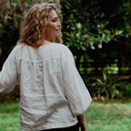Woman wearing a light-colored blouse standing outdoors with greenery in the background