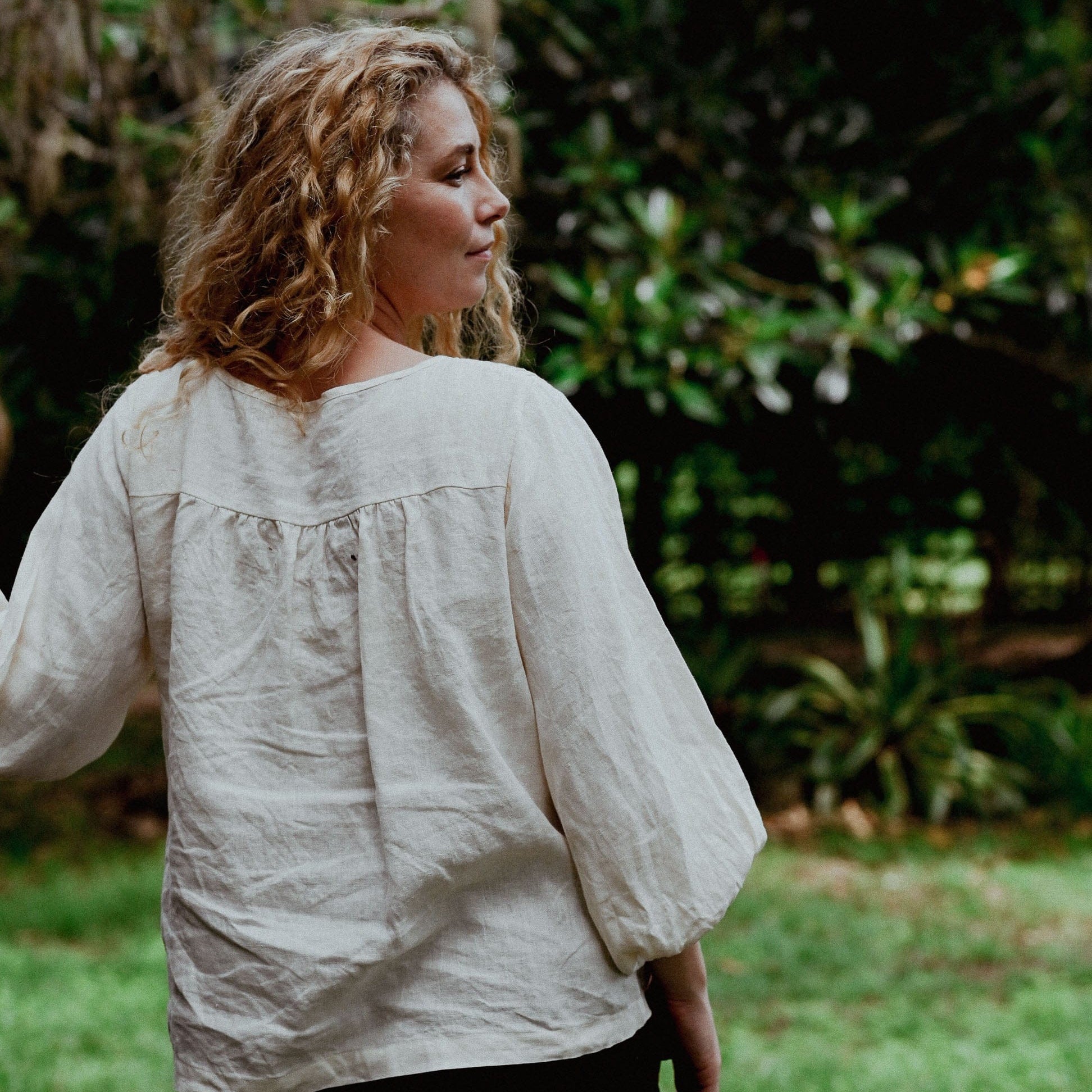 Woman wearing a light-colored blouse standing outdoors with greenery in the background
