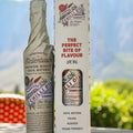 A bottle of Pallmore Chilli Oil next to its white cardboard packaging box, set against a blurred outdoor background of green foliage and blue mountains.