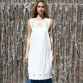 A woman stands in front of a waterfall wall wearing a sleeveless white tunic top with lace detailing at the neckline and eyelet trim at the hem, paired with loose blue jeans and tan mule shoes.