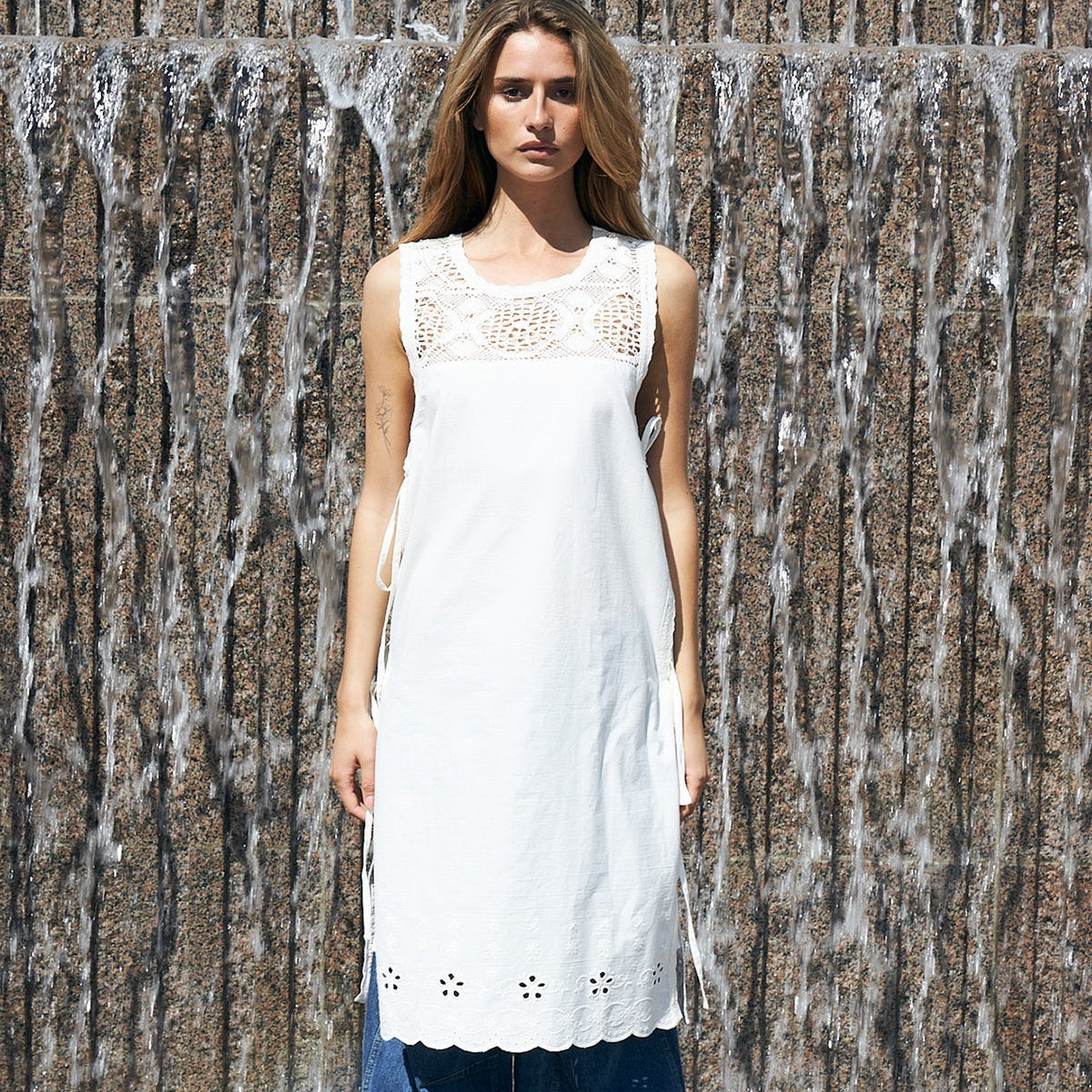 A woman stands in front of a waterfall wall wearing a sleeveless white tunic top with lace detailing at the neckline and eyelet trim at the hem, paired with loose blue jeans and tan mule shoes.