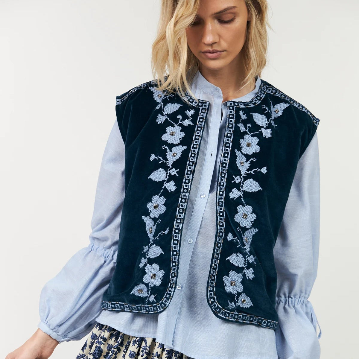 Woman wearing a blue embroidered vest over a light blue shirt with floral patterns on a white background