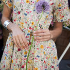 A woman wearing a short-sleeve cream-colored dress with a dense multicolor floral print, holding a purple allium flower.