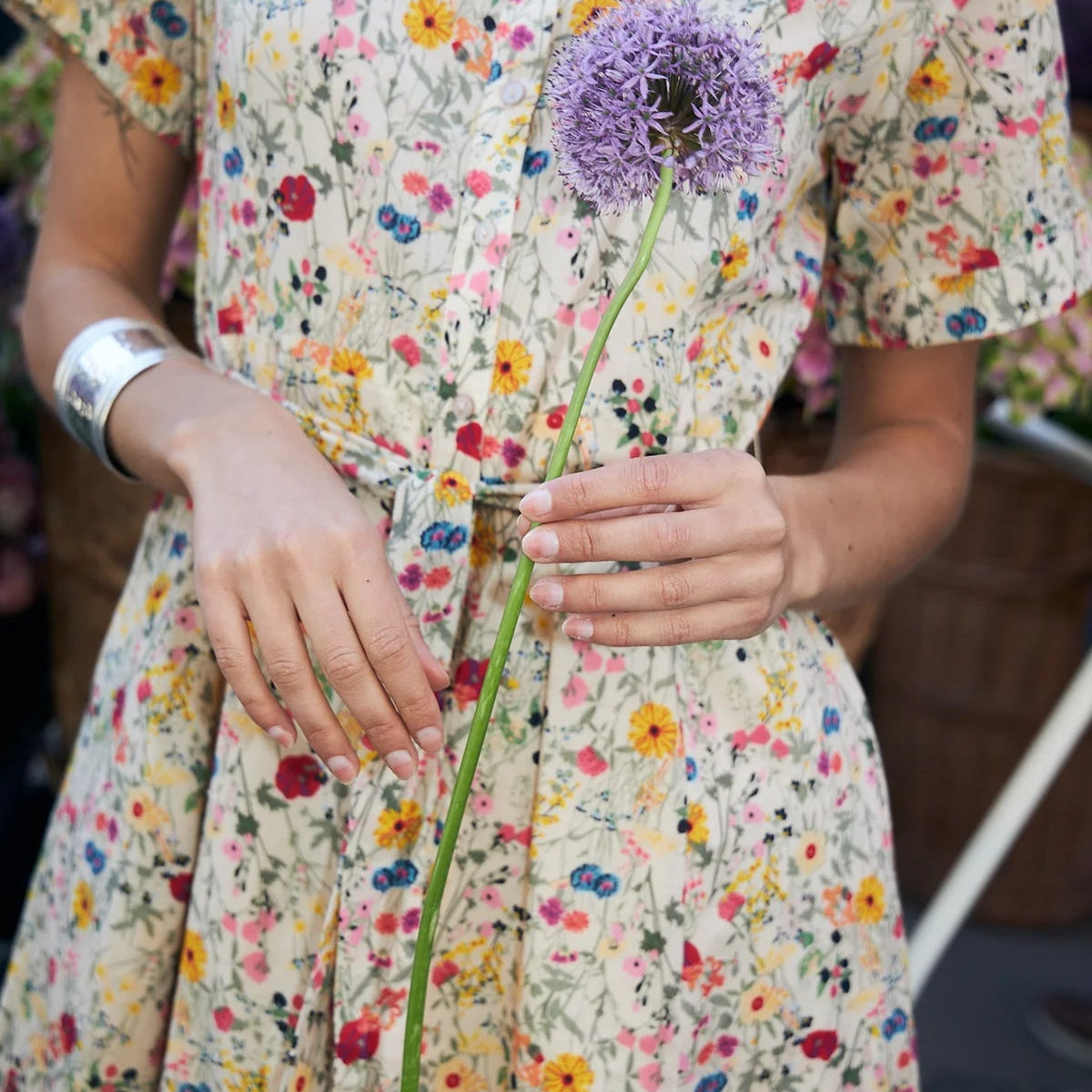 A woman wearing a short-sleeve cream-colored dress with a dense multicolor floral print, holding a purple allium flower.
