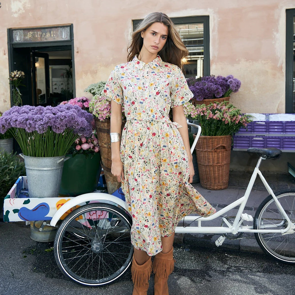 A woman stands outdoors in front of a peach-colored building, wearing a short-sleeve midi dress with a colorful floral print on a cream base and brown fringe ankle boots. A white bicycle and flower baskets are visible beside her.