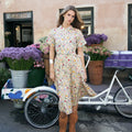 A woman stands outdoors in front of a peach-colored building, wearing a short-sleeve midi dress with a colorful floral print on a cream base and brown fringe ankle boots. A white bicycle and flower baskets are visible beside her.