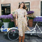 A woman stands outdoors in front of a peach-colored building, wearing a short-sleeve midi dress with a colorful floral print on a cream base and brown fringe ankle boots. A white bicycle and flower baskets are visible beside her.