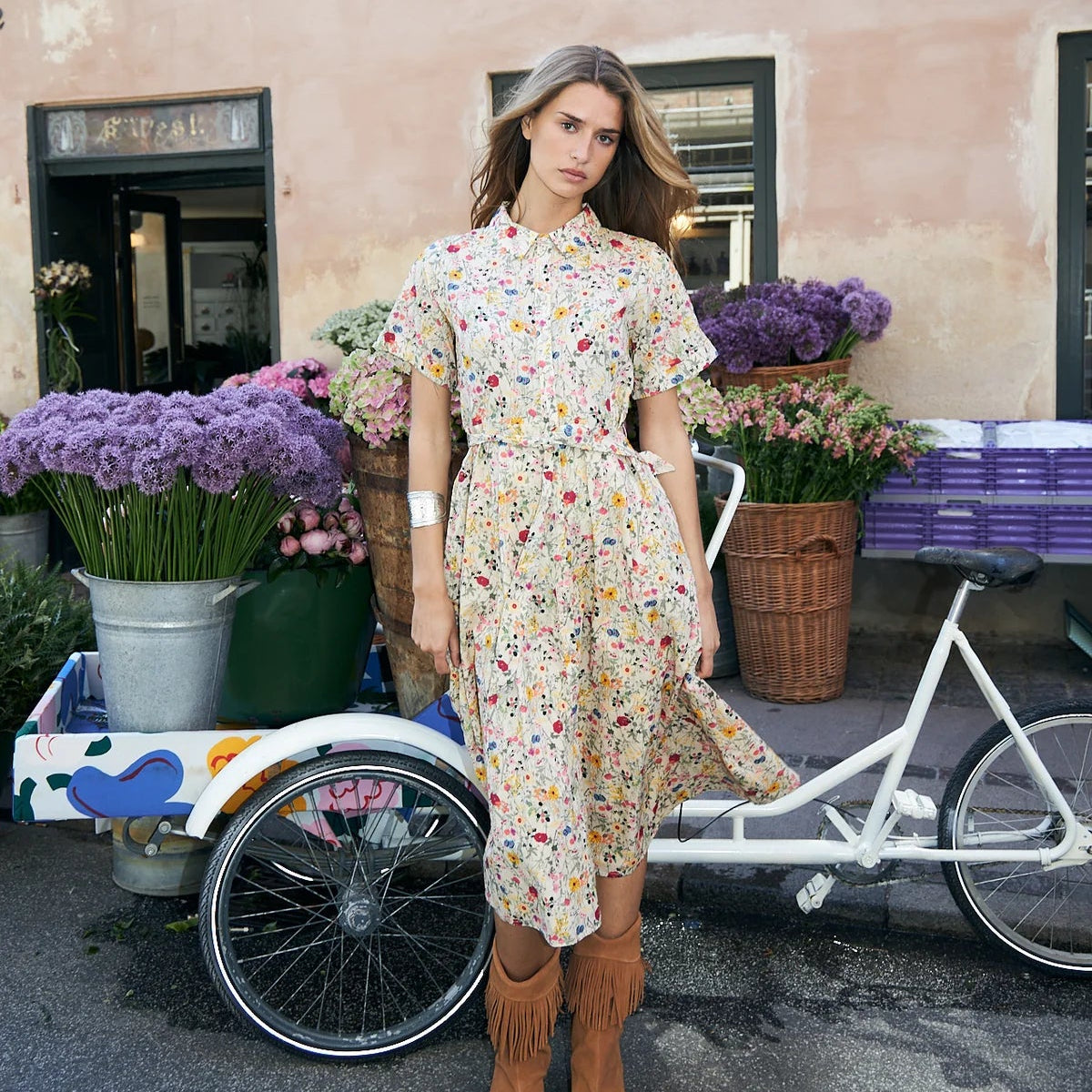 A woman stands outdoors in front of a peach-colored building, wearing a short-sleeve midi dress with a colorful floral print on a cream base and brown fringe ankle boots. A white bicycle and flower baskets are visible beside her.