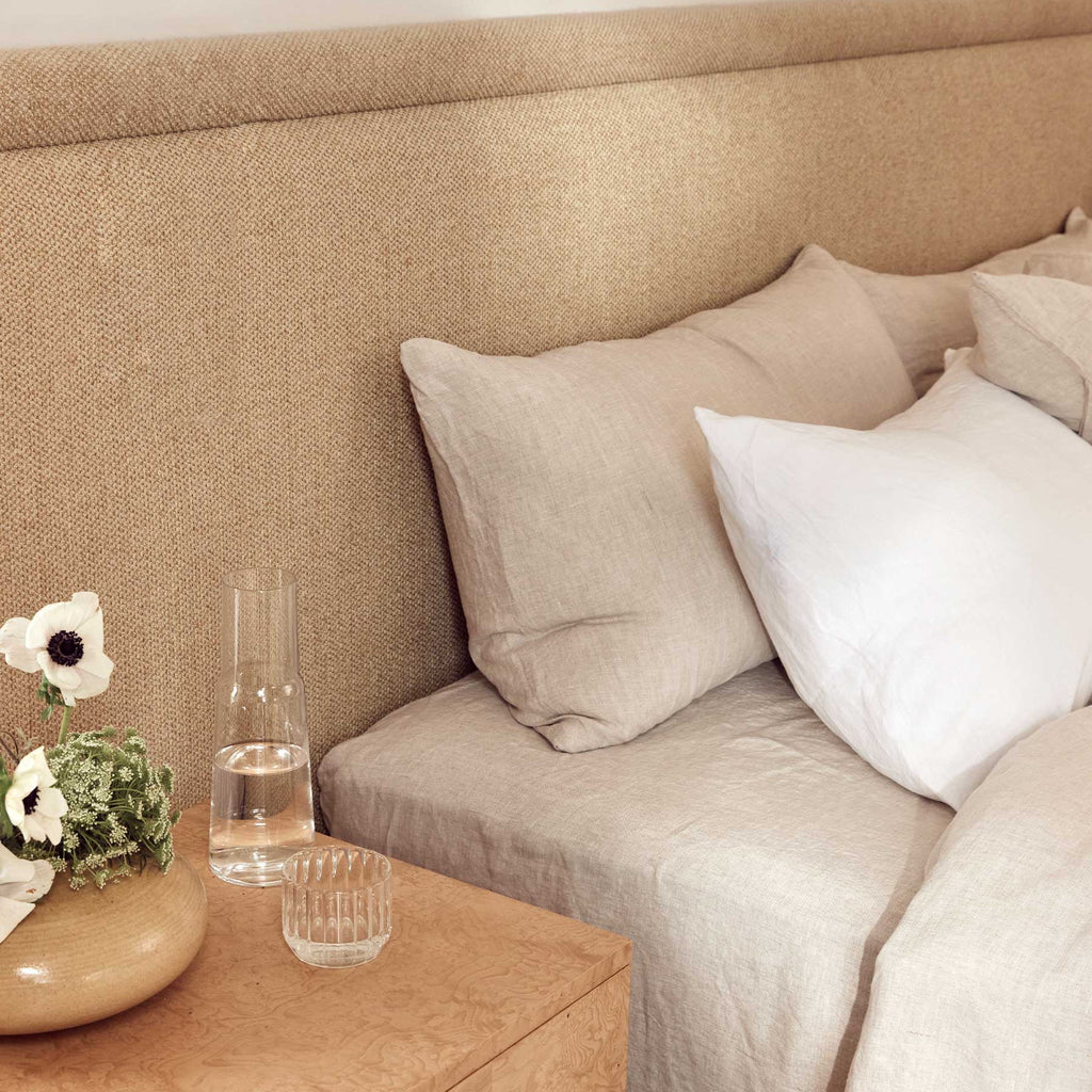 Beige upholstered headboard with pillows and a small table with a vase and glasses.