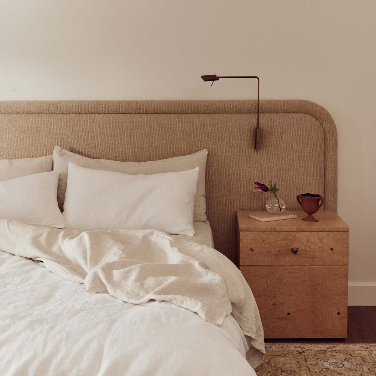 Neatly made bed with white bedding and pillows, nightstand with decorative items, and wall-mounted lamp.