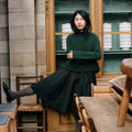 Woman in a green sweater and black skirt standing in a room with wooden furniture and bookshelves.