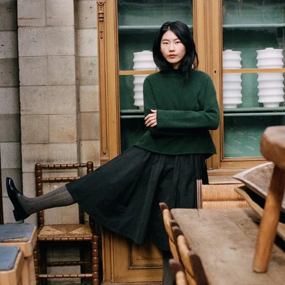 Woman in a green sweater and black skirt standing in a room with wooden furniture and bookshelves.