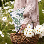Person holding a floral-patterned clutch and a basket of flowers in a field of white flowers.