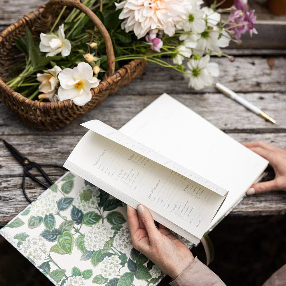 Person holding a book with floral and leaf-patterned notebook on a wooden surface with flowers and a basket.