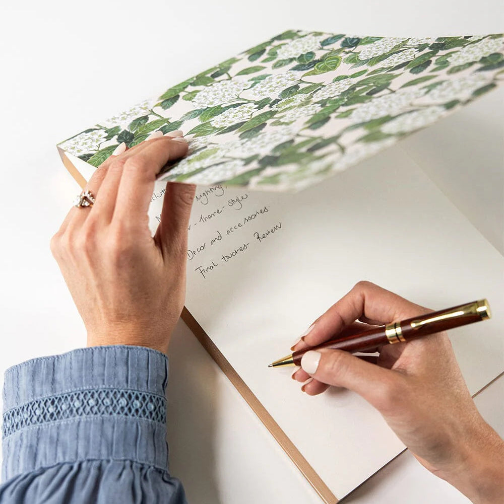 Person writing in a notebook with a floral cover on a white surface