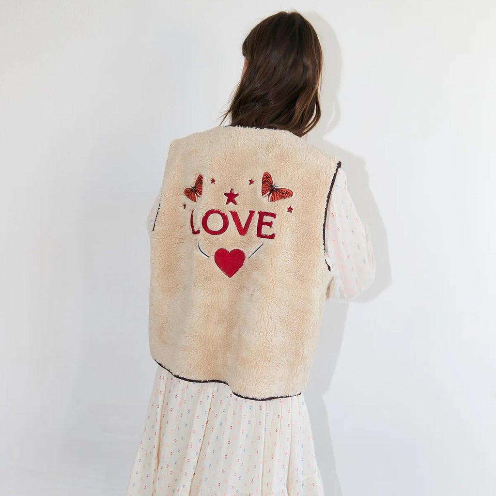 Person wearing a beige fleece vest with 'LOVE' and heart design on the back, standing against a white wall.