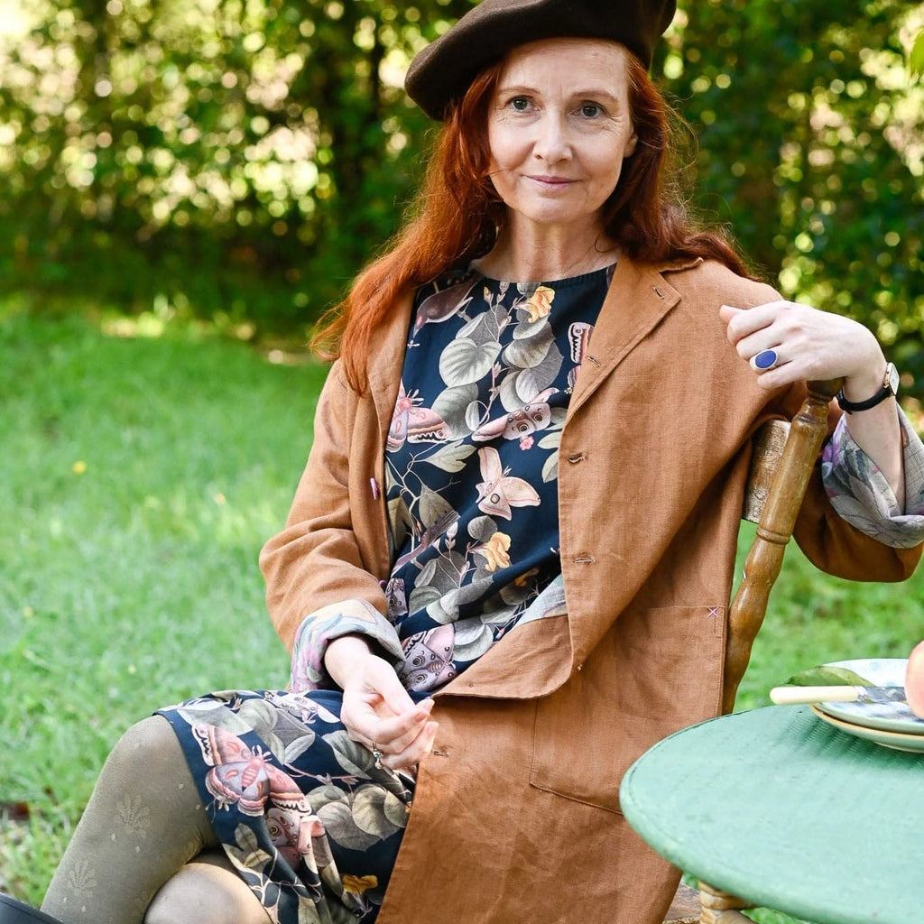 Woman sitting outdoors in a garden wearing a brown coat and beret.