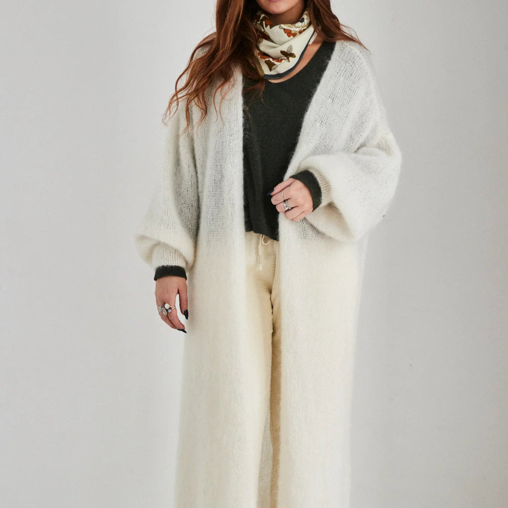 Woman wearing a long cream cardigan over a black sweater and white pants, standing against a white background.