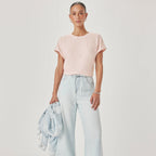 A model stands against a light gray studio background wearing a pale pink short-sleeve top, light blue wide-leg cropped jeans, and metallic flat shoes, while holding a light blue denim jacket.