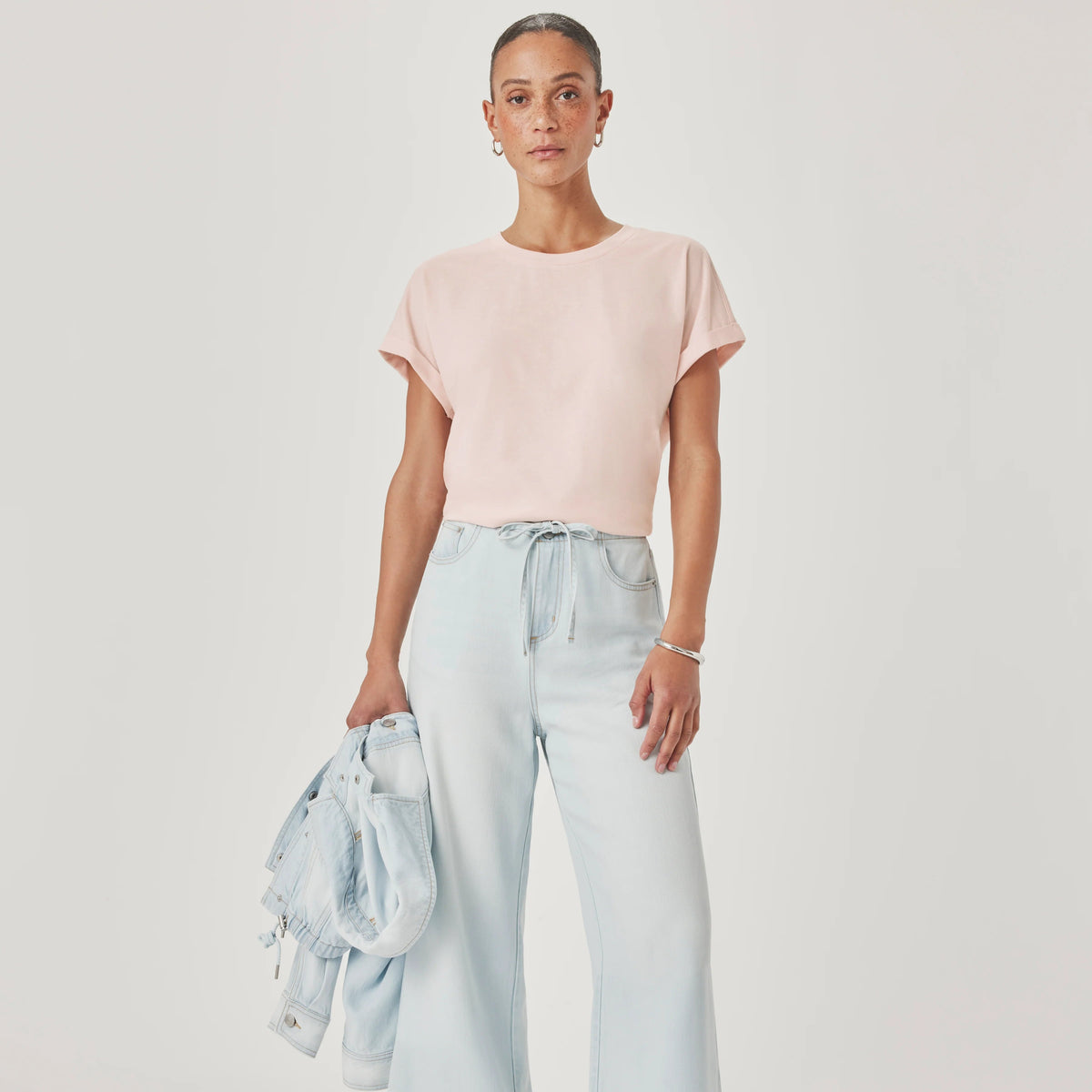 A model stands against a light gray studio background wearing a pale pink short-sleeve top, light blue wide-leg cropped jeans, and metallic flat shoes, while holding a light blue denim jacket.