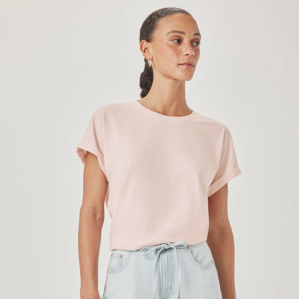 A woman wearing a pale pink short-sleeve crewneck t-shirt and light blue jeans, standing against a plain light gray studio background.