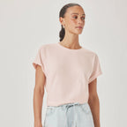A woman wearing a pale pink short-sleeve crewneck t-shirt and light blue jeans, standing against a plain light gray studio background.