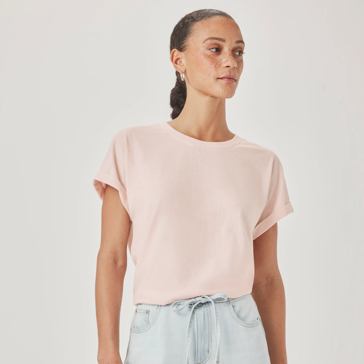 A woman wearing a pale pink short-sleeve crewneck t-shirt and light blue jeans, standing against a plain light gray studio background.