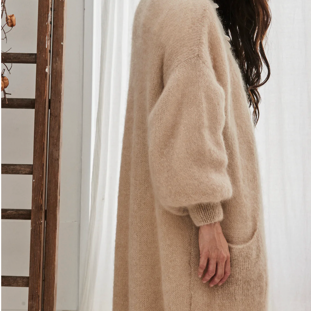 Person wearing a long beige cardigan in a softly lit room with white curtains.