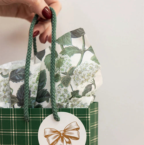 Green plaid gift bag with floral pattern and gold bow label, held by a hand.