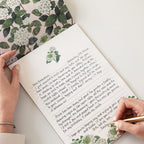 Person writing a letter with floral stationery on a white surface
