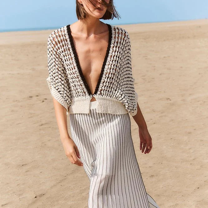 A woman walking barefoot on a sandy beach wearing a cream and black knit cardigan with a deep V-neck and a matching cream and black pinstriped maxi skirt.