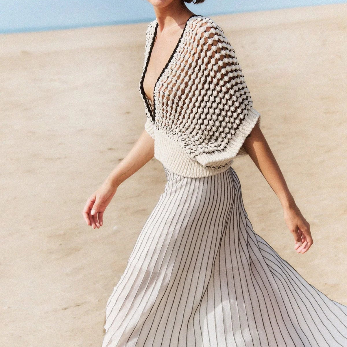 A woman walking on a sandy beach wearing a cream and black striped midi skirt with a chunky knit open-knit vest draped over her shoulders.