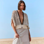 A woman stands in a sandy beach setting wearing a chunky knit beige and black cardigan with a deep V-neck and a cream pleated midi skirt with thin black stripes.